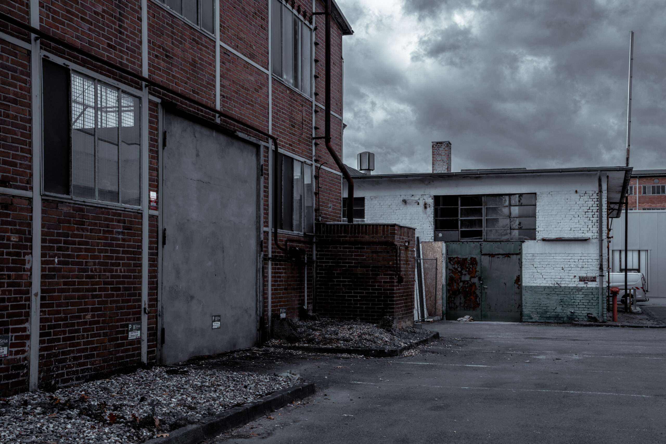 Eine verlassene Industrieanlage mit roten Backsteinmauern und einem alten, weiß gestrichenen Nebengebäude. Die Fassade zeigt Verfallsspuren, mit rostigen Türen, verschmutzten Wänden und bröckelndem Putz. Der Himmel ist dunkel und wolkenverhangen, was der Szene eine triste, vernachlässigte Stimmung verleiht.
