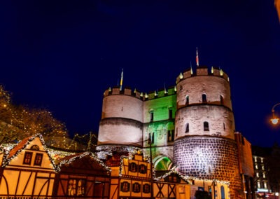Ein historisches Stadttor mit zwei runden Türmen wird nachts stimmungsvoll beleuchtet. Davor stehen festlich geschmückte Fachwerkstände eines Weihnachtsmarkts mit Lichterketten, vor einem tiefblauen Nachthim