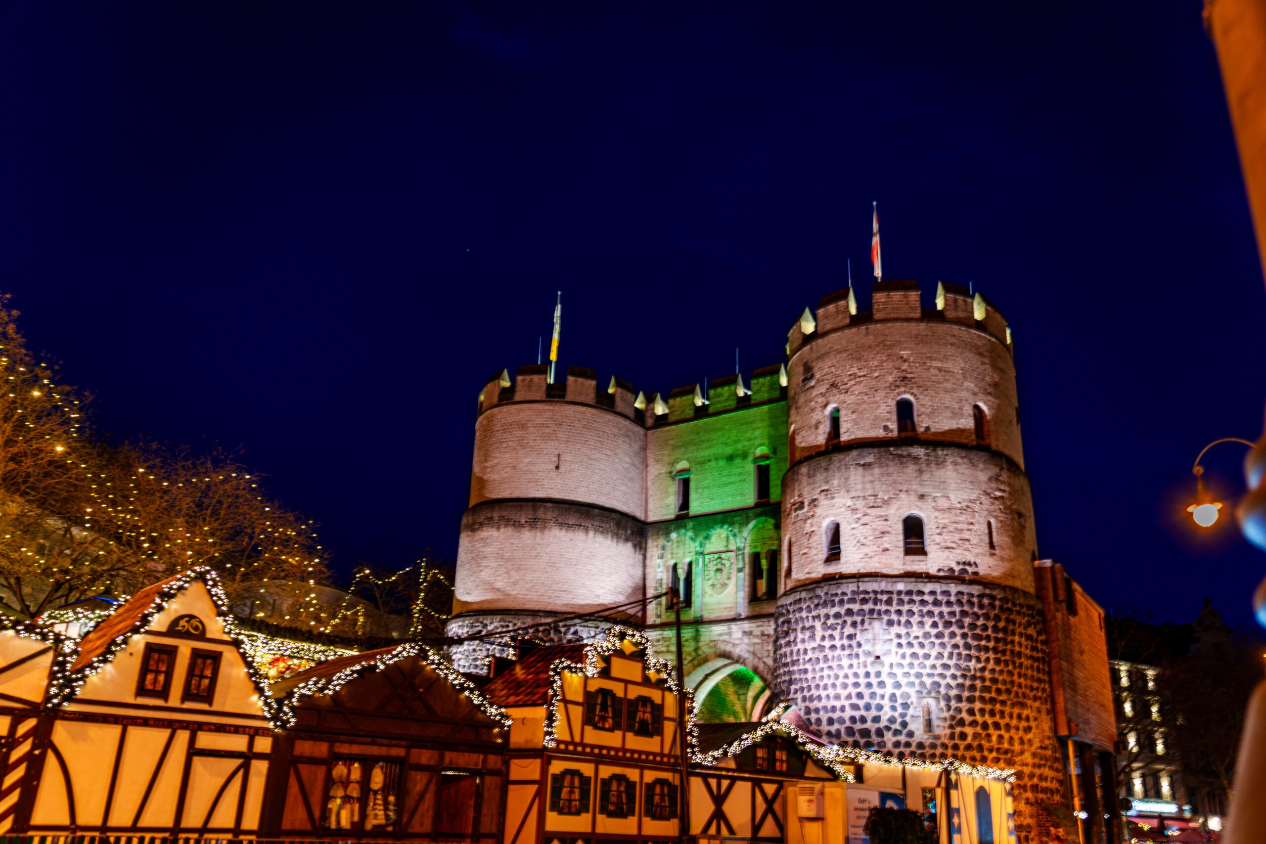 Ein historisches Stadttor mit zwei runden Türmen wird nachts stimmungsvoll beleuchtet. Davor stehen festlich geschmückte Fachwerkstände eines Weihnachtsmarkts mit Lichterketten, vor einem tiefblauen Nachthim